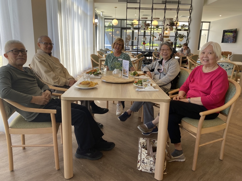 Residents casually sitting around a wooden table in the café smiling. Four residents have a winter lunch in front of them on the table. 