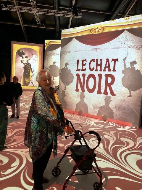 Senior woman with walker standing next to Le Chat Noir artwork on a digital screen.